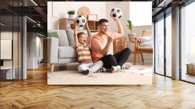 Young man and his little son cheering for football team with soccer balls at home Wall mural