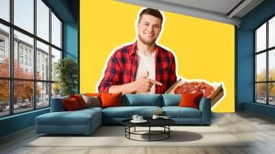 Young man pointing at cardboard box with tasty pepperoni pizza on yellow background Wall mural