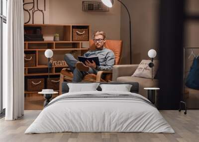 Young man reading book while sitting in armchair at home Wall mural