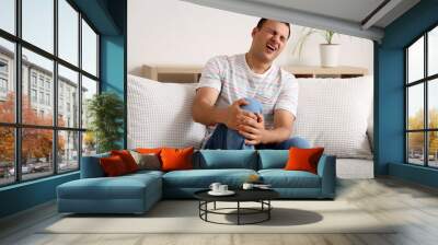 Young man suffering from pain in his knee on sofa at home Wall mural