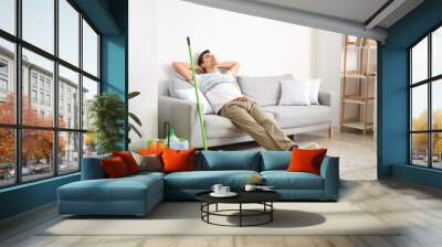 Young man with cleaning supplies relaxing in living room Wall mural