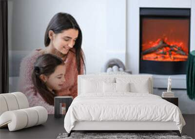 young mother with daughter reading book near fireplace at home Wall mural