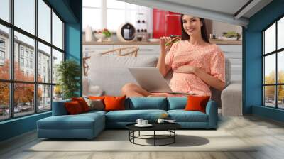 Young pregnant woman with tasty sandwich and laptop in kitchen Wall mural