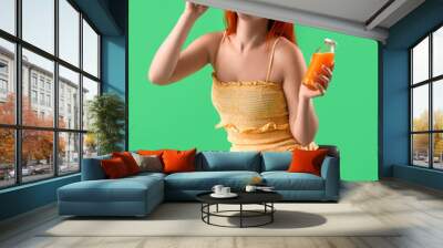 Young redhead woman with bottle of juice eating tasty sandwich on green background Wall mural