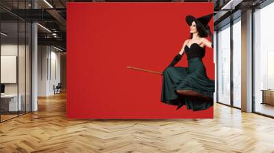 Young witch flying on broom against red background with space for text Wall mural