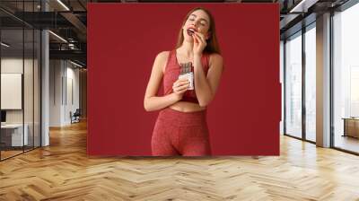 Young woman eating sweet chocolate bar on red background Wall mural