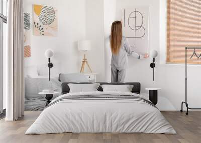 Young woman hanging poster on light wall in bedroom Wall mural