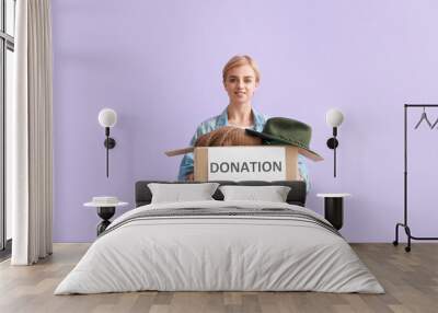 Young woman holding box with donation clothes on lilac background Wall mural