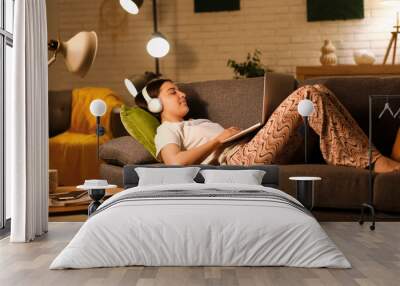 Young woman in pajamas and headphones using laptop on cozy sofa in living room at night Wall mural