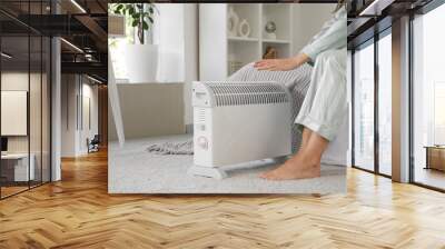 Young woman in pajamas sitting on bed near electric heater in bedroom, closeup Wall mural