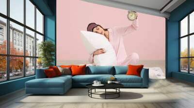 Young woman in pajamas with blanket and pillow holding alarm clock on pink background Wall mural