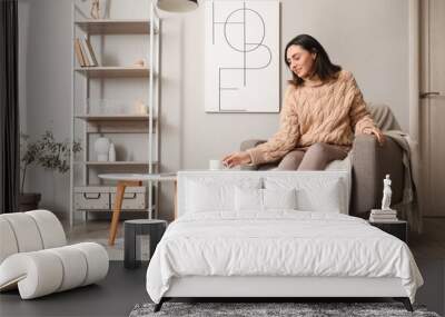 Young woman in soft armchair taking cup of coffee from table at home Wall mural