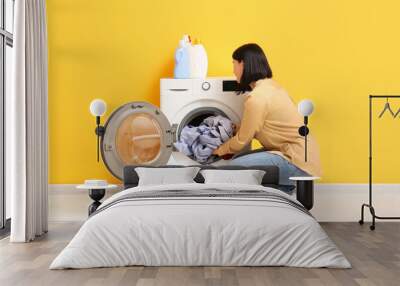 Young woman putting dirty clothes into washing machine on orange background Wall mural