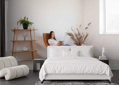 Young woman reading book while sitting in armchair with foot rest in room Wall mural