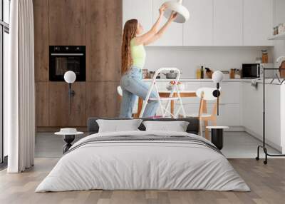 Young woman screwing light bulb into lamp on stepladder in kitchen Wall mural