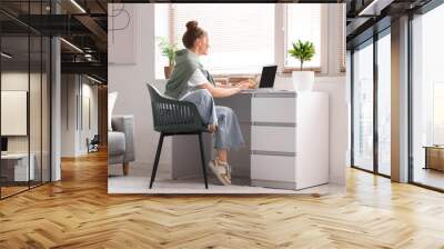 Young woman sitting at workplace and working with laptop in living room Wall mural