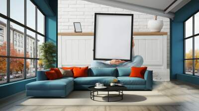 Young woman sitting near chest of drawers and holding blank picture frame in room Wall mural