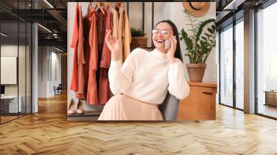 Young woman talking by mobile phone near clothes rack at home Wall mural