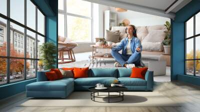 Young woman with headphones meditating on carpet in living room Wall mural