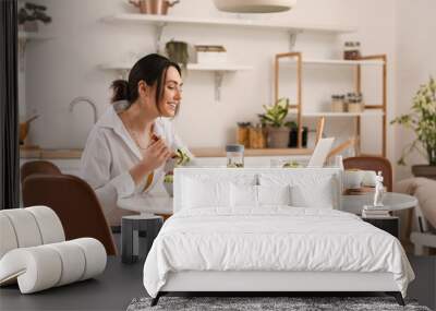 Young woman with laptop eating vegetable salad at table in kitchen Wall mural