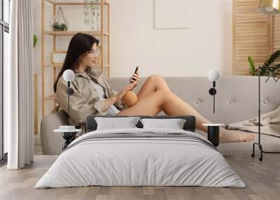 Young woman with mobile phone massaging her leg on sofa at home Wall mural