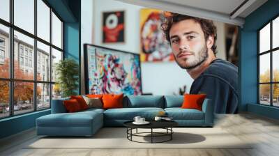 Young Male Designer with Colorful Digital Art. A Caucasian young man with brown wavy hair and light skin, dressed in casual attire, sits in a creative studio with abstract digital artwork on his monit Wall mural