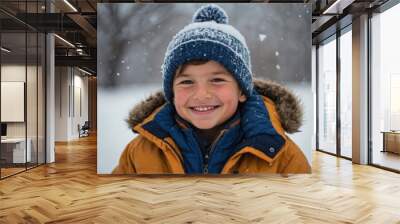 A young boy in a blue jacket and hat is standing in the snow, surrounded by a winter landscape. His warm clothing contrasts with the white, snowy background, capturing the essence of a cold winter day Wall mural