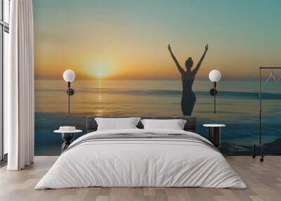 Silhouette of a woman standing on the beach at sunrise with arms raised, facing the ocean Wall mural