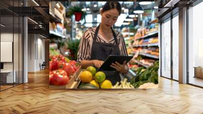 Female shop owner checking inventory lists and managing supplier communications on her tablet in her grocery store, surrounded by organic produce. Wall mural