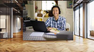 Happy young Asian teenage of a boy wear wireless headset studying online class on laptop computer at home smiling to camera. Wall mural