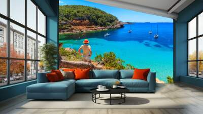 Young woman tourist standing on rock cliff edge and looking at beautiful Cala Salada bay famous for its azure crystal clear sea water, Ibiza island, Spain Wall mural