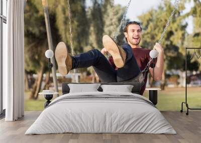 Happy and cheerful young male adult person having fun riding a swing at the park's playground during a sunny summer day. Wall mural