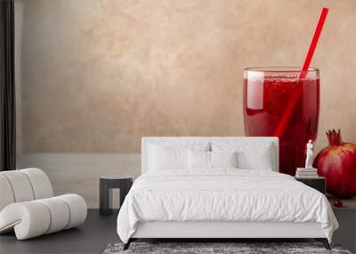 A bright red glass of pomegranate juice, served with a straw and pomegranate seeds scattered on the table, against a neutral backdrop Wall mural