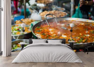 A Thai street food vendor serving bowls of Tom Yum Goong soup to customers, with a large pot of simmering broth and a variety of fresh ingredients on display at a bustling market. Wall mural