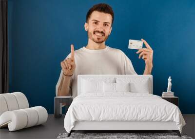 Happy casual man wearing t-shirt posing isolated over blue background holding credit card and pointing with hand and finger to the side. Wall mural