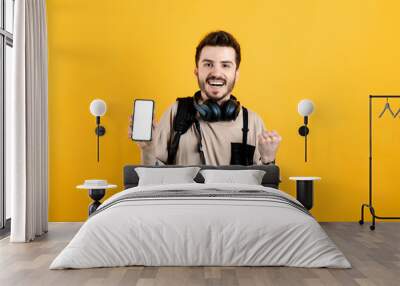 Happy young man wearing beige t-shirt posing isolated over yellow background holding and showing phone with white blank screen to camera. Smiling and screaming for success. Wall mural