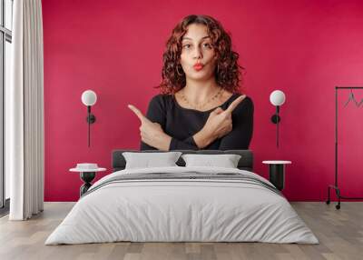 Redhead millennial woman wearing black dress standing isolated over red background crosses arms and points sideways, chooses between two options. Shocked, an incredible choice. Wall mural