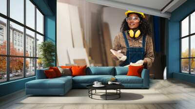 Young attractive black female carpenter wearing safety goggles and hard hat for woodworking in small business workshop. Wood  industry and furniture industry. Wall mural