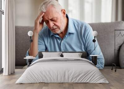 Senior man sits on a couch at home suffering from a headache or migraine with copy space. Wall mural