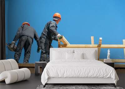 Repair of a wooden roof outdoors on a summer day against the background of blue sky and clouds. A carpenter in special clothes and with a tool installs beams and wood boards. Wall mural