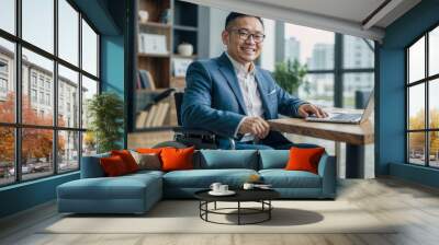 Disabled Asian businessman in a suit and wheelchair, smiling and working with a laptop in a modern office Wall mural