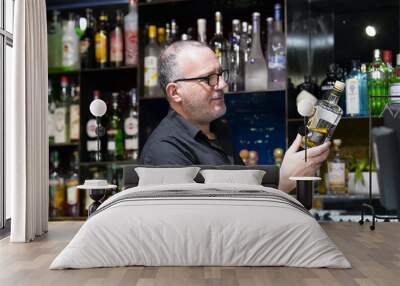 Elderly bartender is holding a bottle of whiskey. The barman examines a bottle of whiskey. The bartender pours whiskey to the client in the hotel bar. The concept of service.  Wall mural