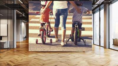 Mother goes pedestrian crossing with children on bicycles. A woman with son and daughter crossing the road in the city. Back view. Tone Wall mural