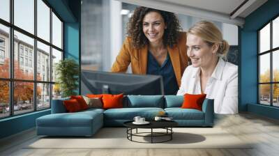 Two women are sitting at a computer, smiling and working together. Concept of collaboration and teamwork, as the women are focused on their task and enjoying each other's company Wall mural