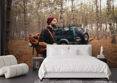 Millennial man hipster with beard wearing backpack, holding axe and firewood in forest with car on background. Wall mural