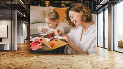 Mother and daughter making Valentine's day cards using color paper, scissors and pencil, sitting by the table in cozy room Wall mural