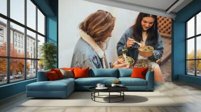 Two young women female friends sitting outdoors eating takeaway food, laughing and having fun. Food delivery and takeout. Wall mural