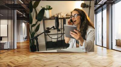 Young woman blogger with long black hair recording online podcast using her laptop, headphones and professional microphone in a studio. Wall mural