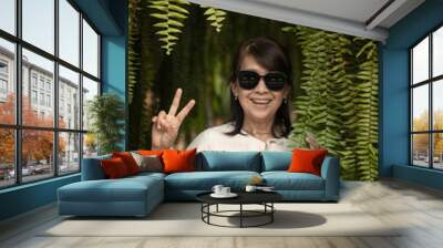 An Asian woman in casual dress clothing wearing black glasses is smiling and raising her hand in the midst of the nature of green fern leaves. Wall mural