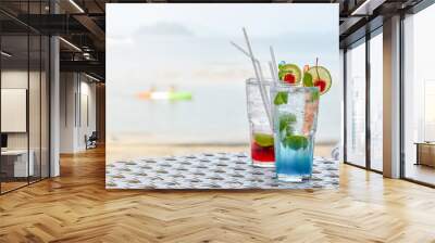 Cold fancy colored summer drinks with lemon slice, mint and cherries on a beach side table. Cold refreshment drinks with blurry beach scenery background. Wall mural